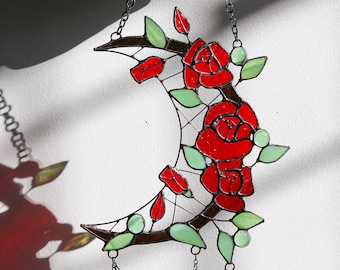 May include: A stained glass crescent moon with red roses and green leaves hanging from a chain. The moon is decorated with a vine and has three chains with green leaves hanging from the bottom.