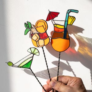 Könnte beinhalten: Cocktaildekorationen aus Buntglas in leuchtenden Farben. Das Set enthält einen Martini und zwei tropische Getränke mit Orangenscheiben, einem roten Regenschirm und einem Strohhalm. Die Getränke werden von einem dünnen, gedrehten Drahtstiel gehalten.