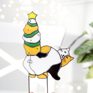 May include: A stained glass cat with a black and white body and yellow accents is holding a Christmas tree with a yellow star on top. The tree is decorated with green, white, and red ornaments.