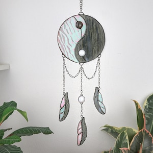 May include: A stained glass yin yang dream catcher with a light blue and gray design. The dream catcher has three feathers hanging from it, two are light blue and gray and one is pink and gray.