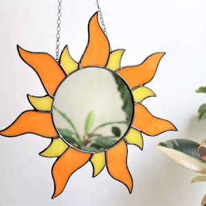 May include: A stained glass sun mirror with orange and yellow glass pieces. The mirror is round and has a black metal frame. The sun is hanging from a chain.