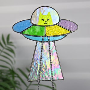 May include: A stained glass UFO with a green cat inside. The UFO is made of iridescent glass and has a blue, green, yellow, and purple design. The UFO is hanging from a string.