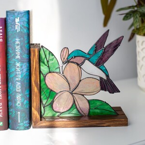 Pu&ograve; includere: Reggilibri in vetro colorato con colibr&igrave; e fiore. Il reggilibri ha una base e una cornice in legno, con un colorato design di colibr&igrave; e fiore. Il colibr&igrave; ha accenti turchesi, bianchi e viola. Il fiore &egrave; rosa e bianco con foglie verdi.