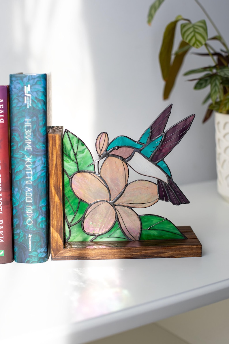 Pu&ograve; includere: Reggilibri in vetro colorato con colibr&igrave; e fiore. Il reggilibri ha una base e una cornice in legno, con un colorato design di colibr&igrave; e fiore. Il colibr&igrave; ha accenti turchesi, bianchi e viola. Il fiore &egrave; rosa e bianco con foglie verdi.