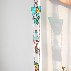 May include: A stained glass hanging mobile with three glass bottles, each containing different colored mushrooms. The bottles are suspended from a chain and are connected to each other by smaller chains. The bottles are a light blue, a light green, and a yellow color.