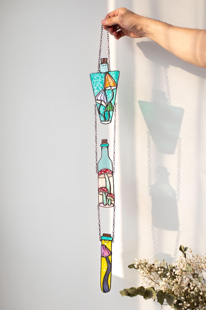 May include: A stained glass hanging mobile with three glass bottles, each containing different colored mushrooms. The bottles are suspended from a chain and are connected to each other by smaller chains. The bottles are a light blue, a light green, and a yellow color.