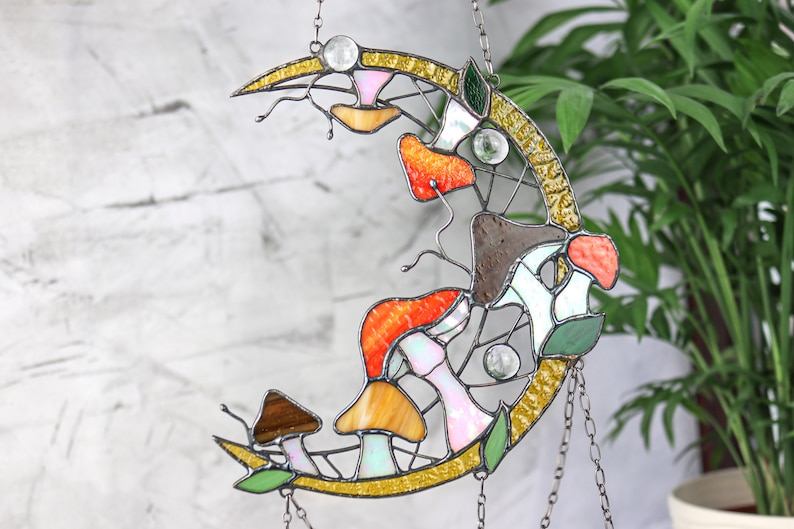 Puede incluir: Un atrapasol de vidrio tintado en forma de luna con un dise&ntilde;o de setas en tonos naranja, amarillo, marr&oacute;n y blanco. El atrapasol est&aacute; colgado de una cadena.