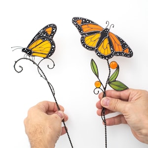 May include: Two decorative butterfly ornaments. One butterfly is perched on a twisted wire stem, the other on a stem with leaves and orange spheres. The butterflies have orange and yellow wings with black details.