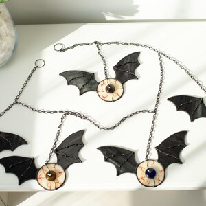 May include: A set of five hanging ornaments featuring black stained glass bat wings attached to circular glass eyeballs with coloured irises and red vein details. Each ornament is connected by a dark metal chain for hanging display purposes.