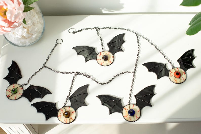 May include: A set of five hanging ornaments featuring black stained glass bat wings attached to circular glass eyeballs with coloured irises and red vein details. Each ornament is connected by a dark metal chain for hanging display purposes.