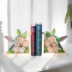 Pu&ograve; includere: Reggilibri in legno con un design di colibr&igrave; e fiori. I reggilibri sono dipinti con colori verdi, rosa e viola. Due libri sono posizionati tra i reggilibri su una superficie bianca.