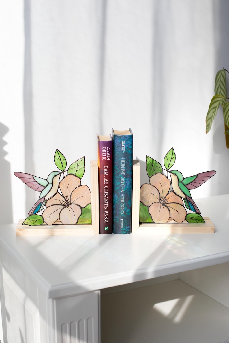 Pu&ograve; includere: Reggilibri in legno con un design di colibr&igrave; e fiori. I reggilibri sono dipinti con colori verdi, rosa e viola. Due libri sono posizionati tra i reggilibri su una superficie bianca.