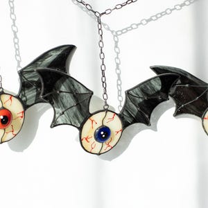 May include: A stained glass hanging ornament featuring three bat-like figures with dark grey, translucent wings. Each bat has a central eyeball design with a white sclera, red veins, and a prominent blue or red iris, suspended by a thin metal chain.