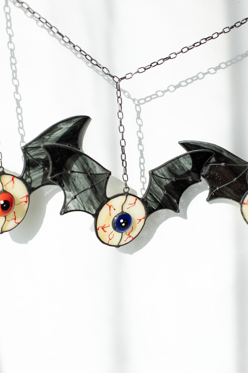 May include: A stained glass hanging ornament featuring three bat-like figures with dark grey, translucent wings. Each bat has a central eyeball design with a white sclera, red veins, and a prominent blue or red iris, suspended by a thin metal chain.