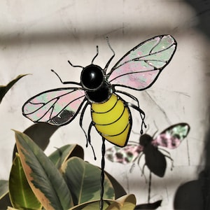 Op de afbeelding: Een glas-in-lood-bij met roze en groene vleugels en een geel lichaam. De bij zit op een metalen steel en is omringd door groene bladeren.