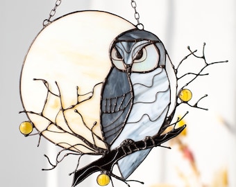 May include: A stained glass window hanging with a gray owl perched on a branch with yellow berries. The owl is in front of a white and yellow stained glass moon.