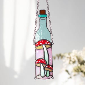 May include: A stained glass bottle with a teal top and a pink background. The bottle contains three red and white mushrooms with green stems.