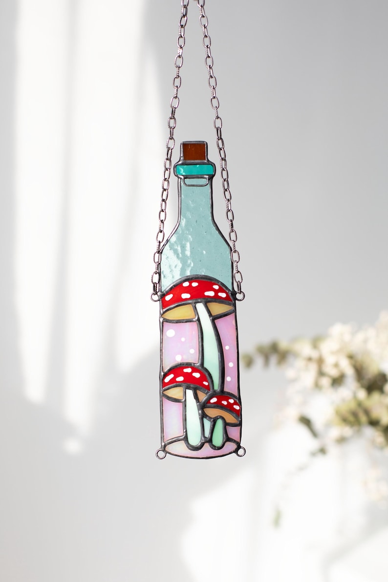 May include: A stained glass bottle with a teal top and a pink background. The bottle contains three red and white mushrooms with green stems.