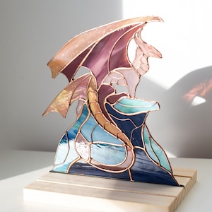 Op de afbeelding: Gebrandschilderde glazen drakenbeeld met een koperkleurig frame. De draak heeft paarse, blauwe en gouden tinten, gezeten op een blauwe en turquoise basis. De sculptuur is gemonteerd op een natuurlijke houten basis.