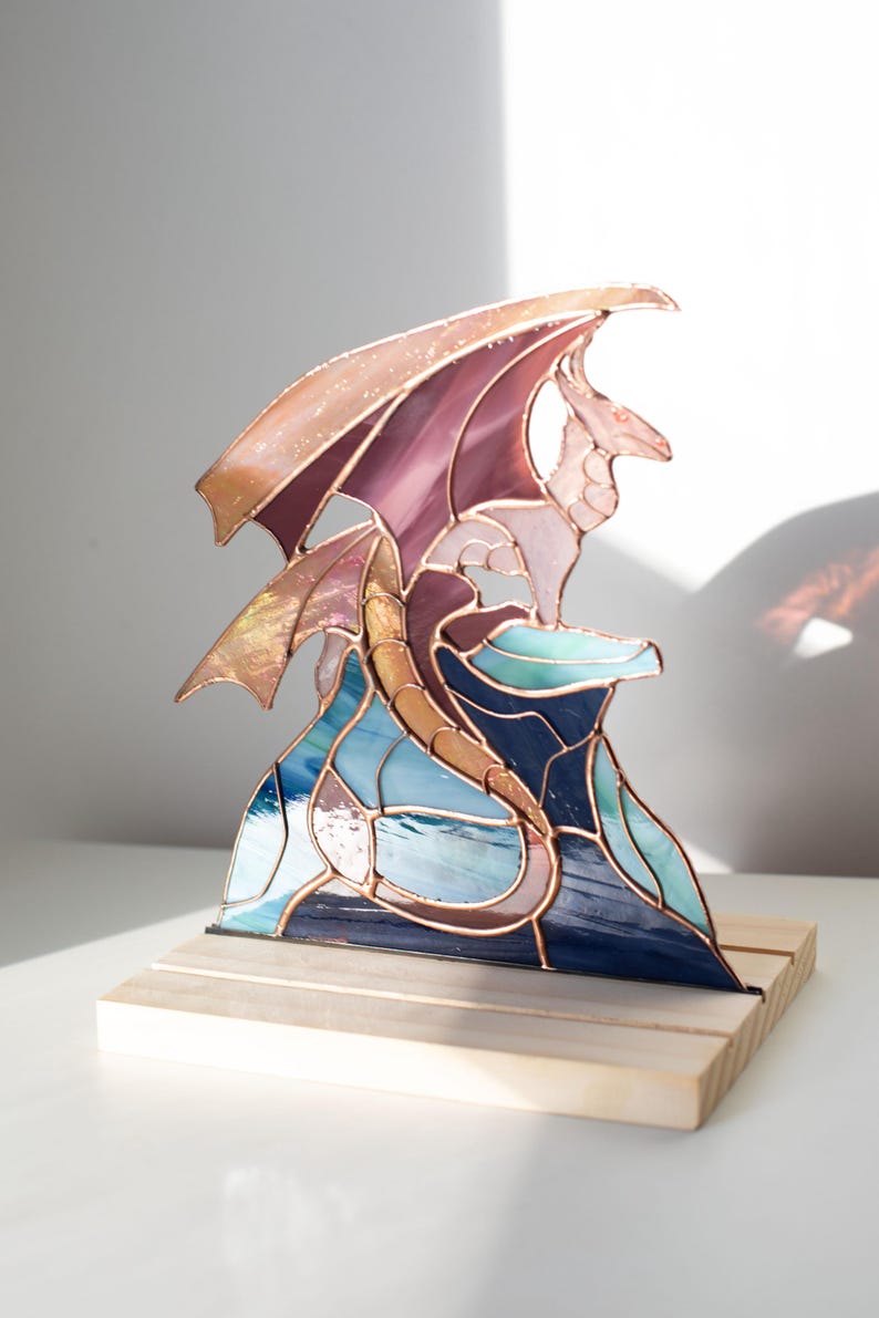 Op de afbeelding: Gebrandschilderde glazen drakenbeeld met een koperkleurig frame. De draak heeft paarse, blauwe en gouden tinten, gezeten op een blauwe en turquoise basis. De sculptuur is gemonteerd op een natuurlijke houten basis.