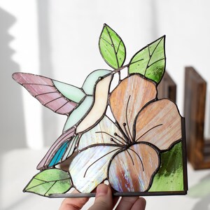 Pu&ograve; includere: Opera d'arte in vetro colorato con colibr&igrave; e fiore. Il colibr&igrave; ha ali e corpo rosa, blu e verdi. Il fiore &egrave; color pesca e bianco con foglie verdi. L'opera d'arte &egrave; tenuta da una mano.
