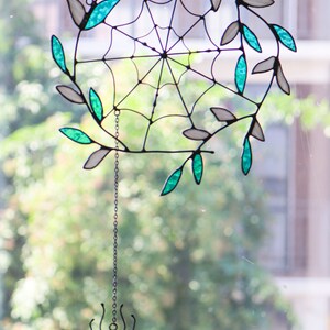 May include: A stained glass spider web with teal and white leaves hanging from a chain with a small stained glass spider at the bottom.