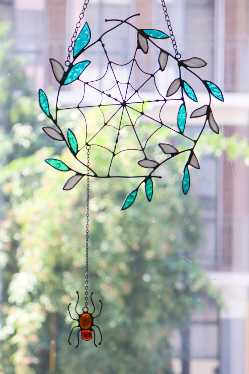 May include: A stained glass spider web with teal and white leaves hanging from a chain with a small stained glass spider at the bottom.