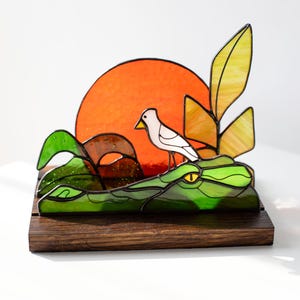 Op de afbeelding: Gebrandschilderde glazen sculptuur met een vogel op een groene alligator, met een oranje zon op de achtergrond. De alligator en het gebladerte zijn groen, de vogel is wit. Gemonteerd op een donkere houten basis.