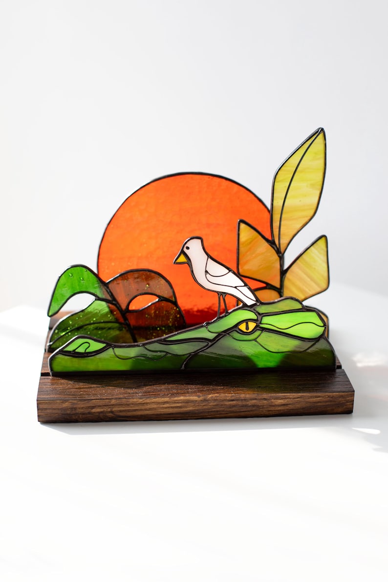 Op de afbeelding: Gebrandschilderde glazen sculptuur met een vogel op een groene alligator, met een oranje zon op de achtergrond. De alligator en het gebladerte zijn groen, de vogel is wit. Gemonteerd op een donkere houten basis.