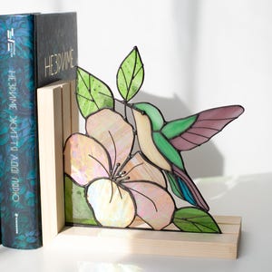 Pu&ograve; includere: Reggilibri in vetro colorato con un design di colibr&igrave; e fiore. Il design colorato presenta un colibr&igrave; con tonalit&agrave; verdi, viola e crema, vicino a un fiore rosa e bianco. Il reggilibri &egrave; posto su una base di legno.