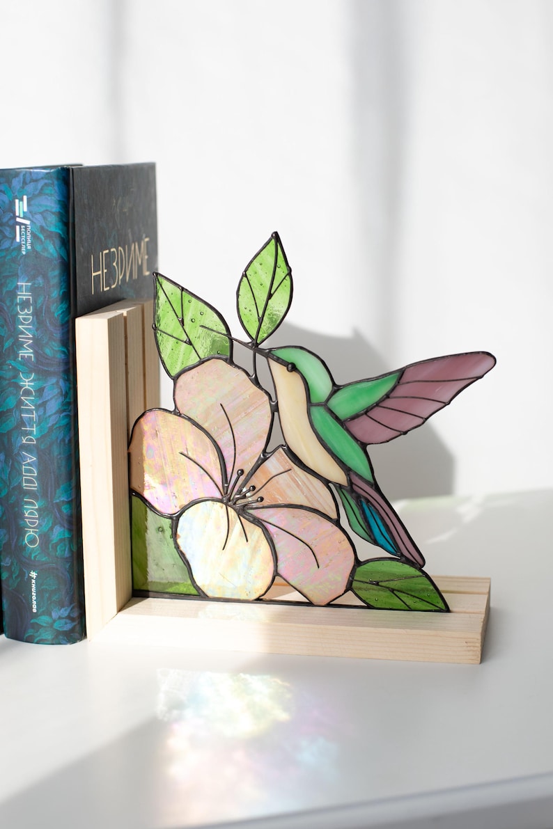 Pu&ograve; includere: Reggilibri in vetro colorato con un design di colibr&igrave; e fiore. Il design colorato presenta un colibr&igrave; con tonalit&agrave; verdi, viola e crema, vicino a un fiore rosa e bianco. Il reggilibri &egrave; posto su una base di legno.