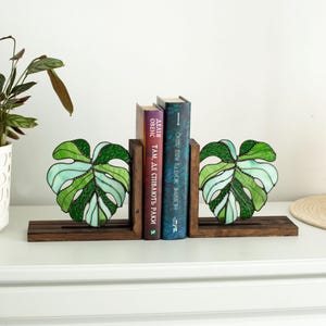 K&ouml;nnte beinhalten: Dekorative Buchst&uuml;tzen aus Holz mit Glasmalerei-Monstera-Bl&auml;ttern in Gr&uuml;n und Wei&szlig;. Die Buchst&uuml;tzen halten zwei B&uuml;cher mit farbigen Buchr&uuml;cken. Die Buchst&uuml;tzen stehen auf einer dunkelbraunen Holzbasis.