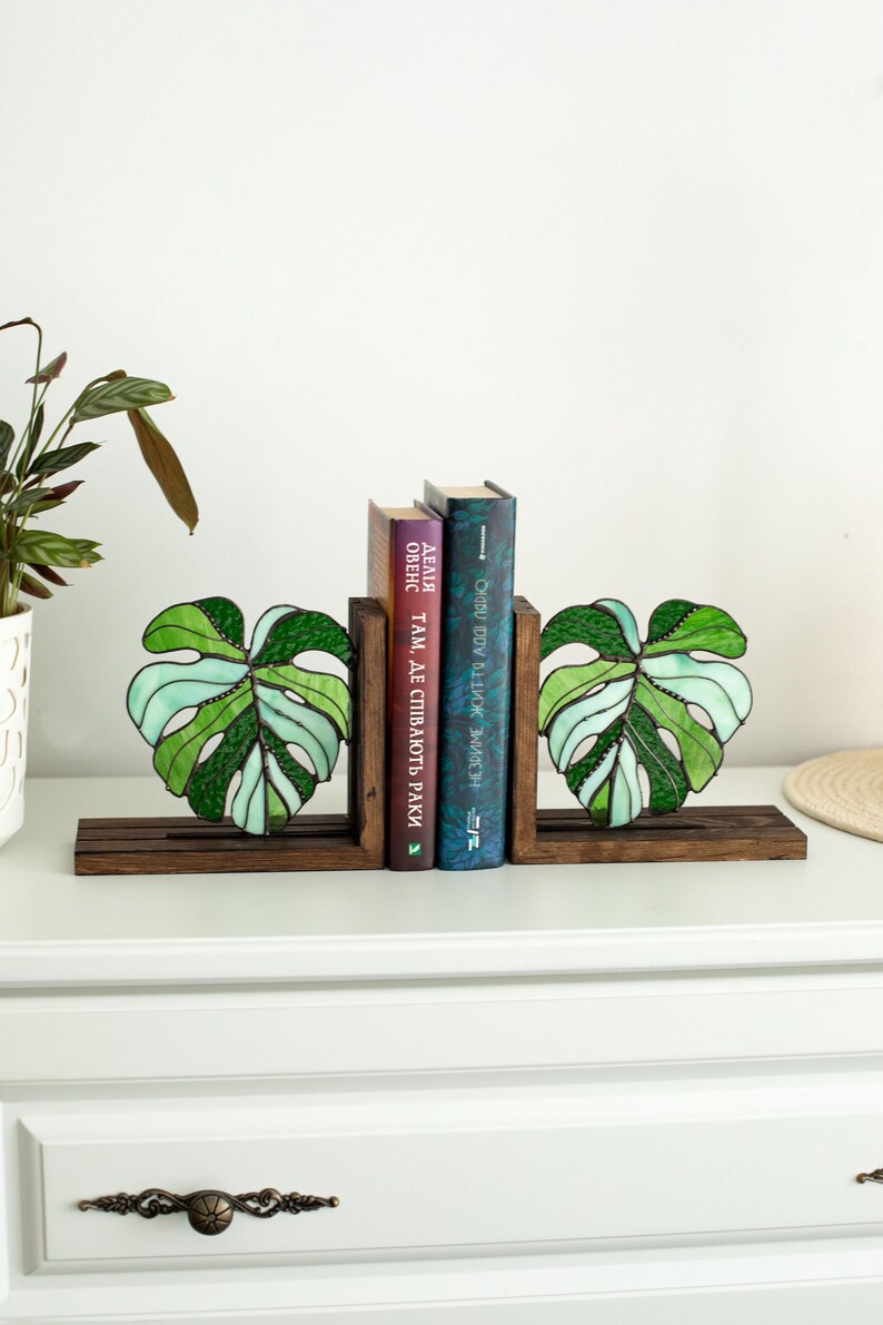 K&ouml;nnte beinhalten: Dekorative Buchst&uuml;tzen aus Holz mit Glasmalerei-Monstera-Bl&auml;ttern in Gr&uuml;n und Wei&szlig;. Die Buchst&uuml;tzen halten zwei B&uuml;cher mit farbigen Buchr&uuml;cken. Die Buchst&uuml;tzen stehen auf einer dunkelbraunen Holzbasis.