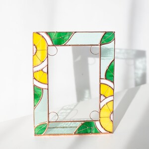 May include: A rectangular stained glass frame with a vibrant design. The frame features yellow, green, white, and light blue glass pieces. The design includes leaf and lemon slice patterns, with copper-colored metal borders and decorative swirls.