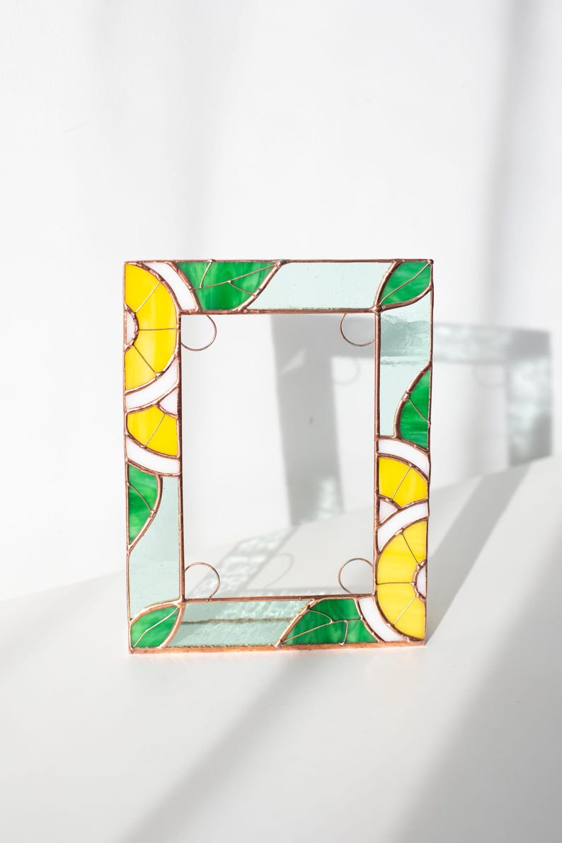 May include: A rectangular stained glass frame with a vibrant design. The frame features yellow, green, white, and light blue glass pieces. The design includes leaf and lemon slice patterns, with copper-colored metal borders and decorative swirls.