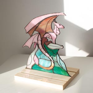 Op de afbeelding: Gebrandschilderde drakenbeeld in roze, oranje en groene tinten. De draak zit op een kleurrijke berg, gemonteerd op een houten voet. De vleugels vertonen een verloop van roze en oranje. Een uniek fantasy kunstwerk.