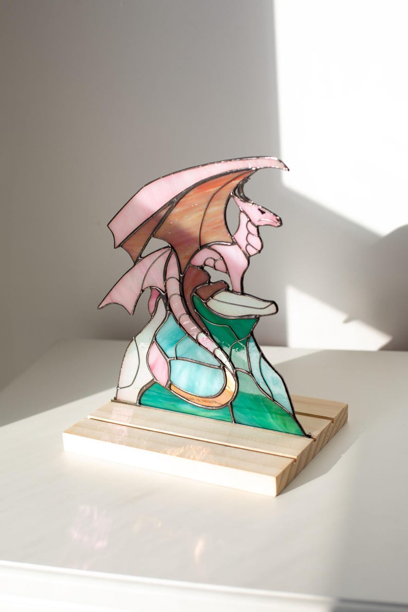 Op de afbeelding: Gebrandschilderde drakenbeeld in roze, oranje en groene tinten. De draak zit op een kleurrijke berg, gemonteerd op een houten voet. De vleugels vertonen een verloop van roze en oranje. Een uniek fantasy kunstwerk.