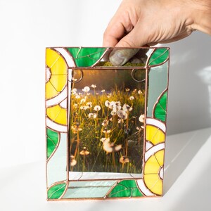 May include: A rectangular stained glass picture frame with a photo of a field of white flowers. The frame has yellow, green, and white geometric designs. The frame is being held up by a hand.