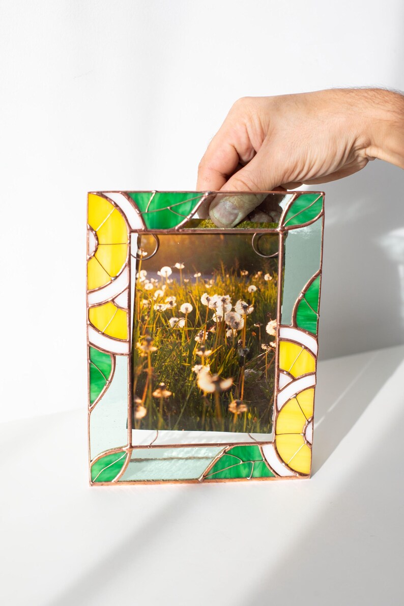 May include: A rectangular stained glass picture frame with a photo of a field of white flowers. The frame has yellow, green, and white geometric designs. The frame is being held up by a hand.