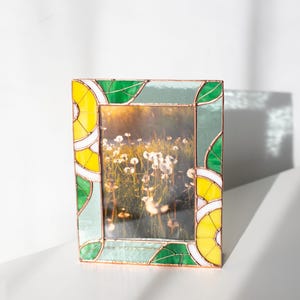May include: A stained glass photo frame with a picture of a field of dandelions. The frame has a copper border and is decorated with green, yellow, and white glass in a leaf and lemon slice design.