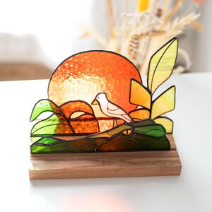 Op de afbeelding: Gebrandschilderd glaskunstwerk met een vogel op een krokodil, met een oranje zon als achtergrond. Het stuk gebruikt groene, gele en oranje kleuren, gemonteerd op een houten voet. Een decoratief item.