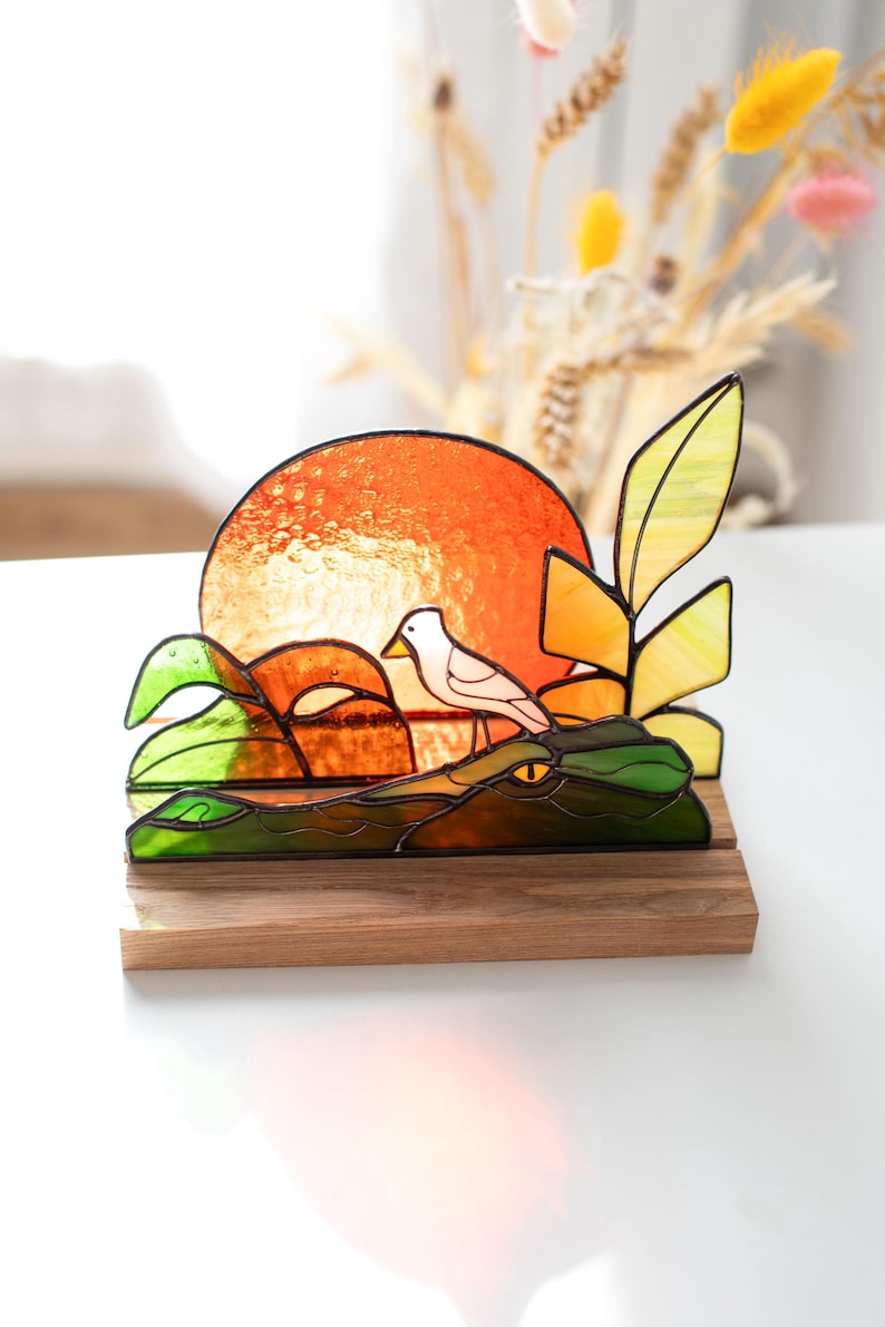 Op de afbeelding: Gebrandschilderd glaskunstwerk met een vogel op een krokodil, met een oranje zon als achtergrond. Het stuk gebruikt groene, gele en oranje kleuren, gemonteerd op een houten voet. Een decoratief item.
