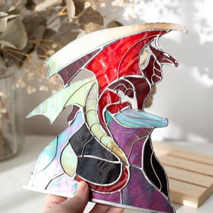 Op de afbeelding: Gebrandschilderde glazen drakenbeeld met rode, witte, paarse, zwarte en iriserende vleugels en lichaam. De draak zit op een paarse en zwarte basis. De sculptuur wordt door een hand vastgehouden.