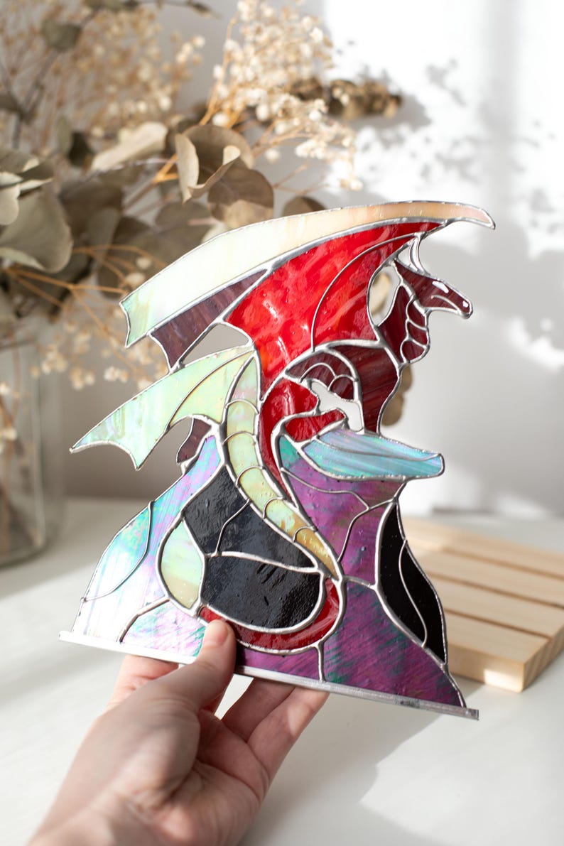 Op de afbeelding: Gebrandschilderde glazen drakenbeeld met rode, witte, paarse, zwarte en iriserende vleugels en lichaam. De draak zit op een paarse en zwarte basis. De sculptuur wordt door een hand vastgehouden.