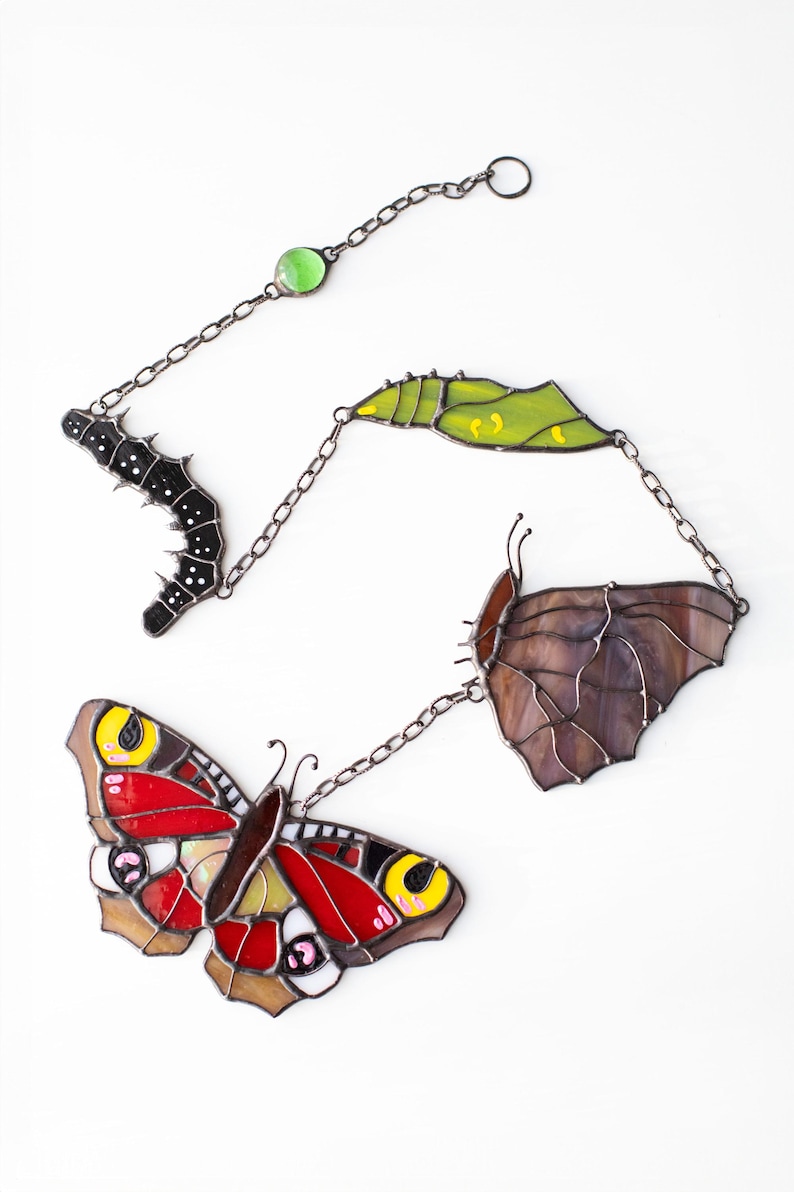 May include: Stained glass butterfly life cycle art piece. The piece includes a caterpillar, chrysalis, and two butterflies in shades of red, yellow, brown, and purple, connected by a chain. The art is set against a white background.