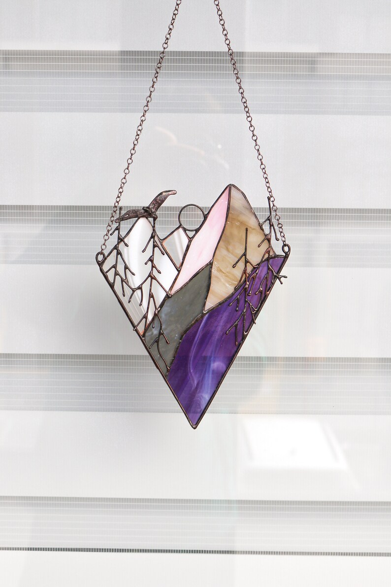 May include: A stained glass window hanging with a copper chain. The window depicts a purple, pink, and brown mountain scene with a bird flying overhead.