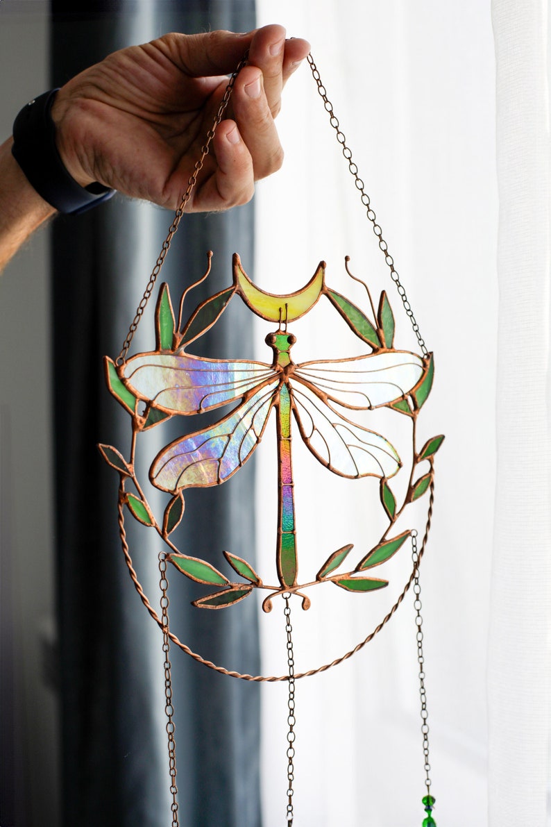 May include: A stained glass dragonfly suncatcher with iridescent wings, a crescent moon, and green leaves. The suncatcher is hanging from a copper chain.