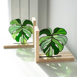 K&ouml;nnte beinhalten: Zwei Buchst&uuml;tzen aus Buntglas in Form von Monstera-Bl&auml;ttern. Jedes Blatt ist eine lebendige Mischung aus hellem und dunklem Gr&uuml;n, eingefasst in einen Holzrahmen. Die Buchst&uuml;tzen sind so konzipiert, dass sie aufrecht stehen.