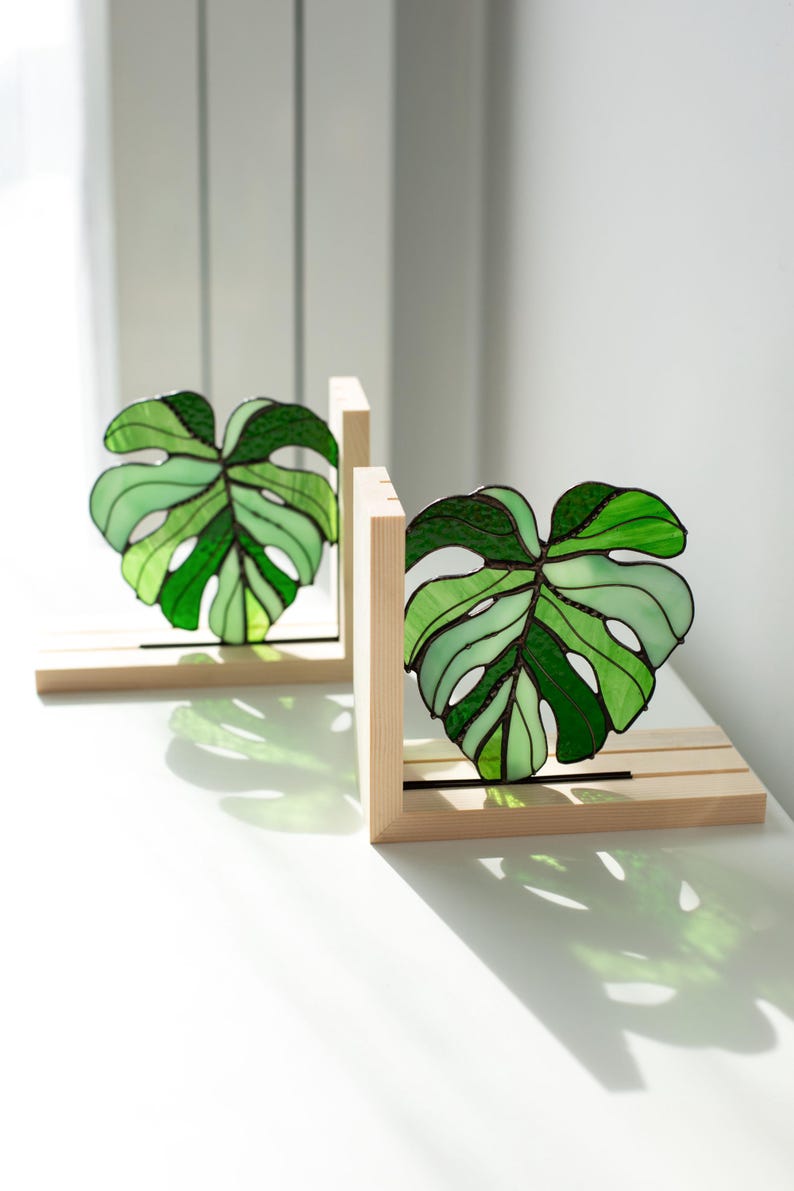 K&ouml;nnte beinhalten: Zwei Buchst&uuml;tzen aus Buntglas in Form von Monstera-Bl&auml;ttern. Jedes Blatt ist eine lebendige Mischung aus hellem und dunklem Gr&uuml;n, eingefasst in einen Holzrahmen. Die Buchst&uuml;tzen sind so konzipiert, dass sie aufrecht stehen.