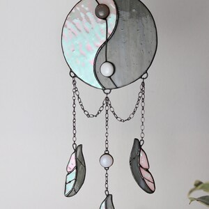 May include: A stained glass yin yang symbol with a gray and iridescent blue and pink design hangs from a chain with three iridescent blue and pink feathers and white beads.